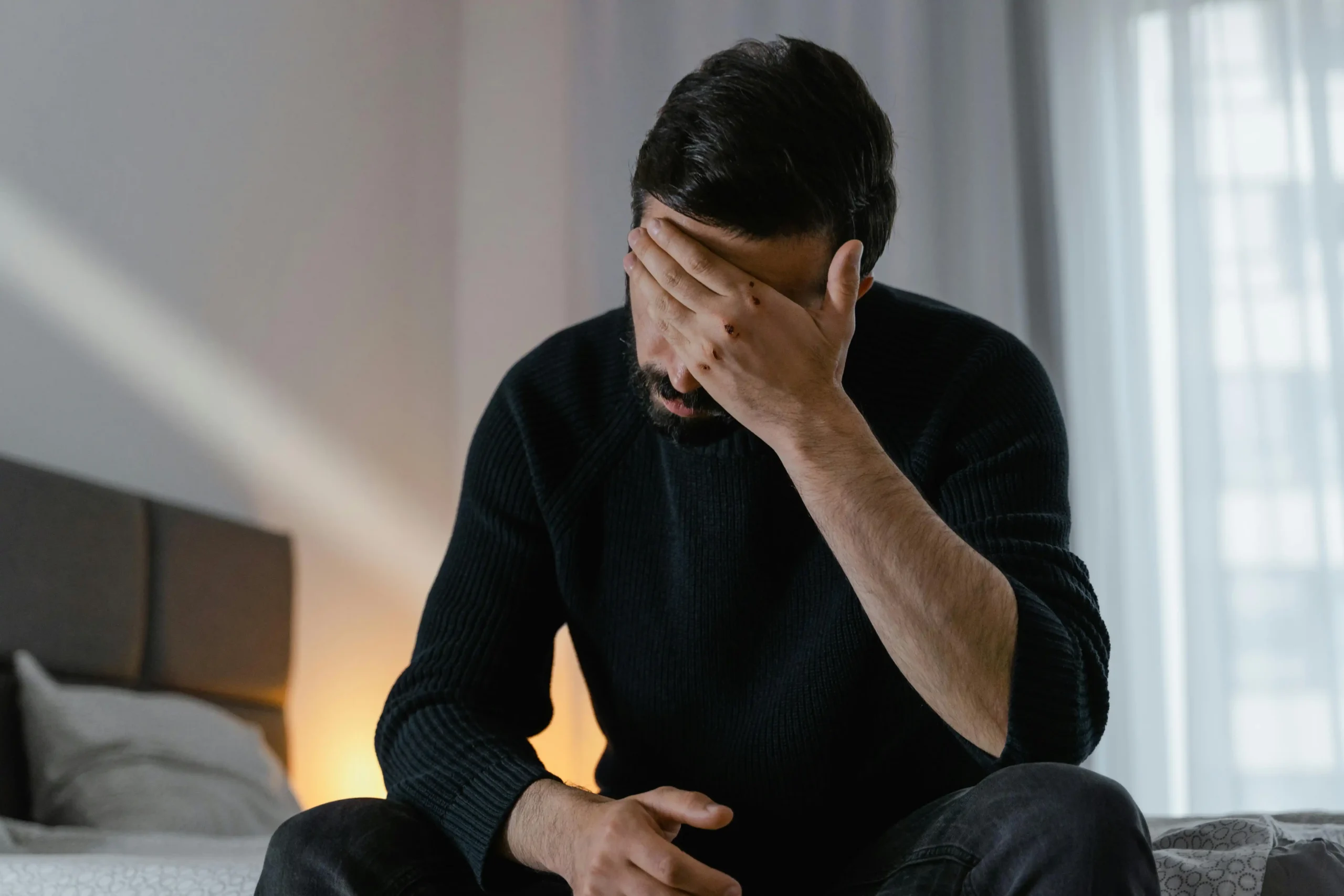 Man in a dark sweater sitting on a bed looking exhausted, illustrating the symptoms of low testosterone that many men dismiss as stress.