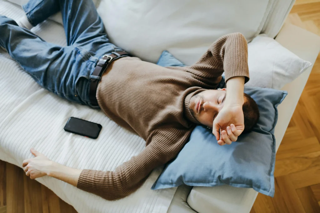 Man lying exhausted on a sofa next to his phone while reviewing documents, illustrating fatigue symptoms often discussed in articles about “What is TRT”.