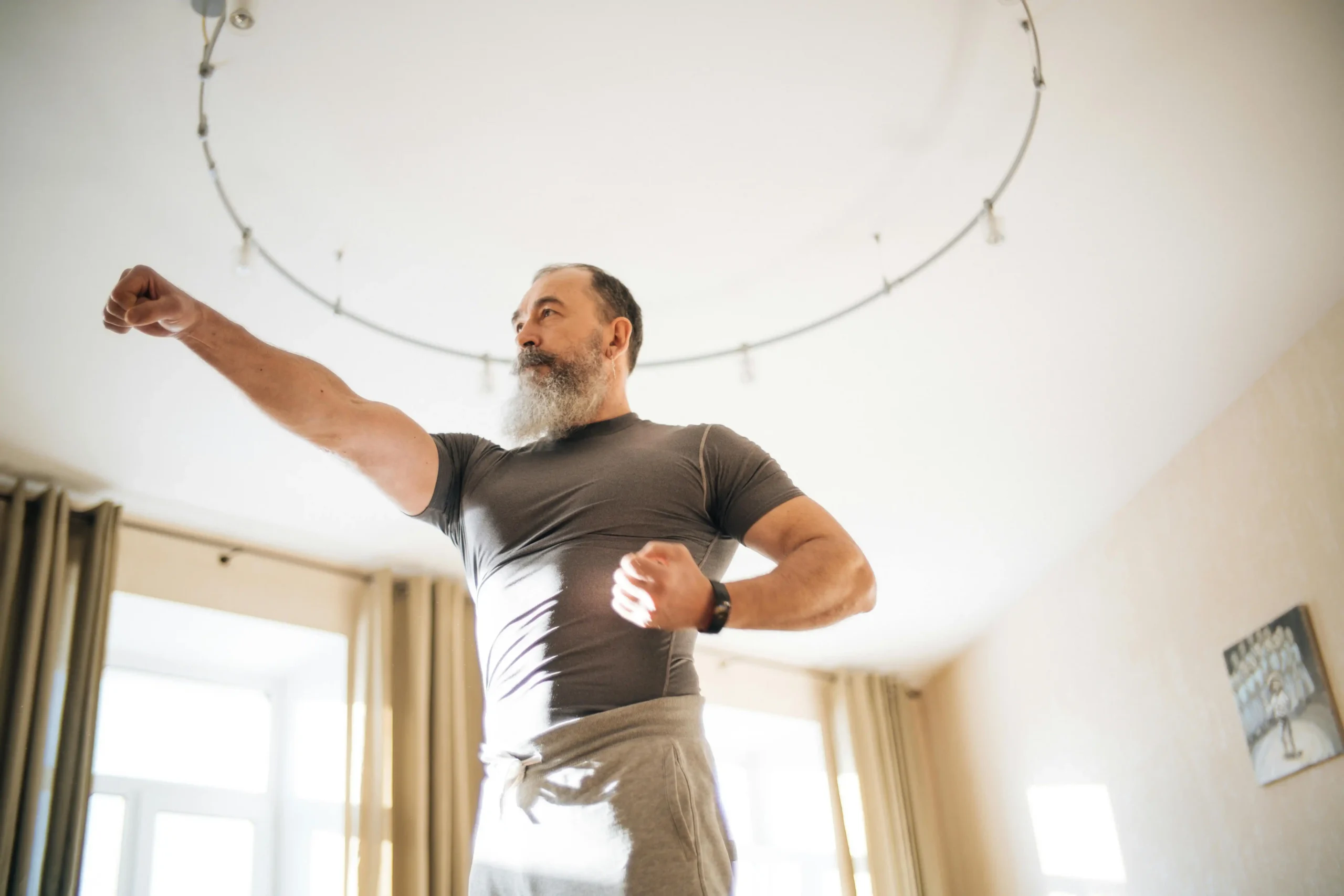 Older bearded man in athletic wear shadow boxing at home, illustrating the energy and vitality men researching testosterone cypionate vs enanthate are often hoping to restore.