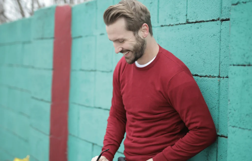 A confident, smiling man in a red sweater leans against a turquoise wall, embodying the potential vitality gains often discussed when comparing natural testosterone vs TRT.