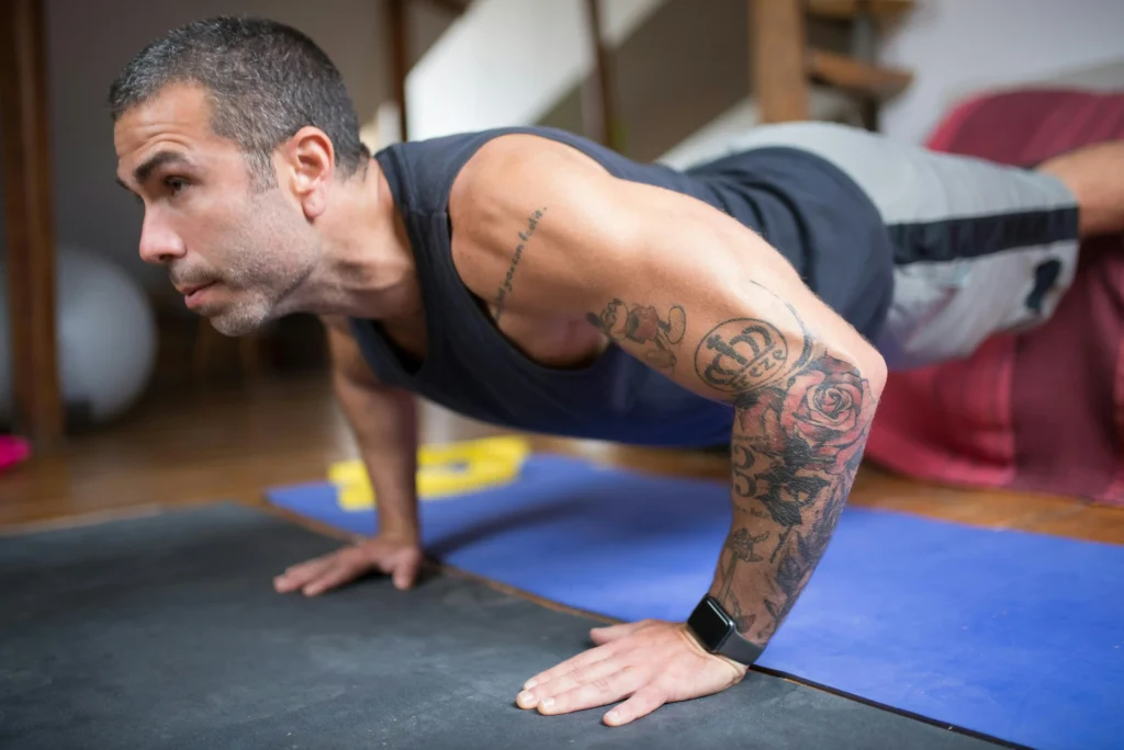 A fit man with arm tattoos performs a push-up on a blue exercise mat at home, representing the physical strength goals often associated with the natural testosterone vs TRT debate.