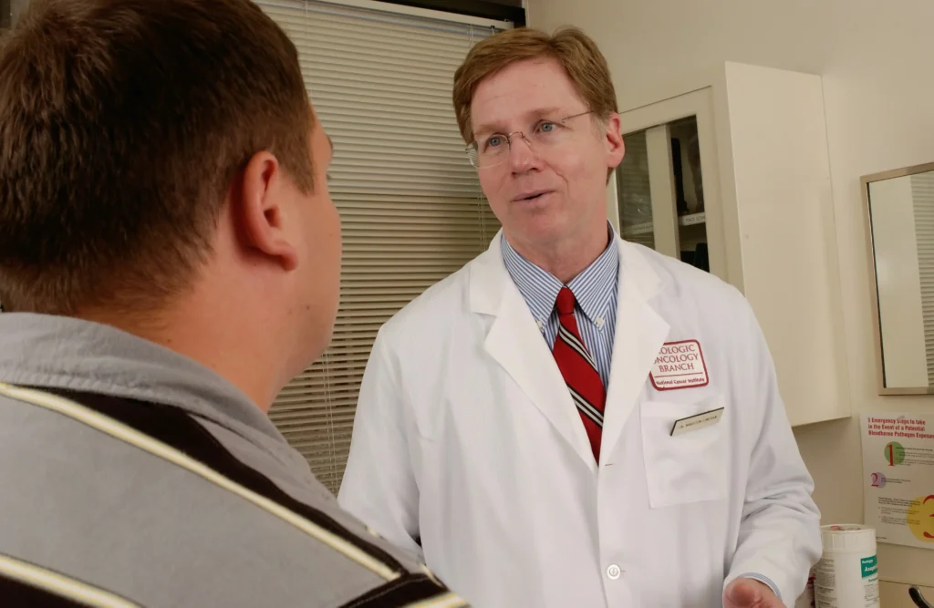 A male doctor in a white lab coat speaks directly to a patient in a clinical setting, emphasizing the importance of professional consultation when discussing testosterone replacement therapy side effects.