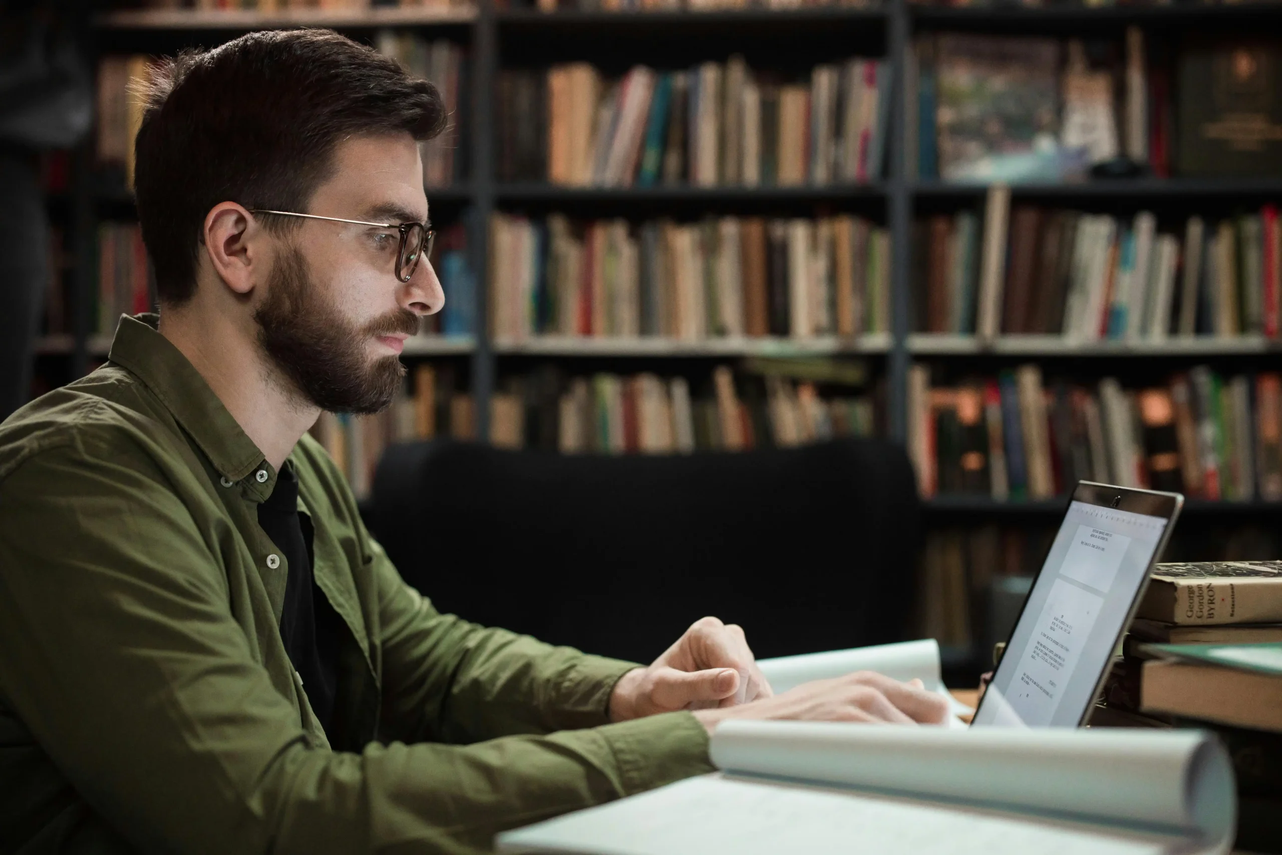 A man in a green shirt and glasses focuses on a laptop in a library researching the long-term risks of TRT.