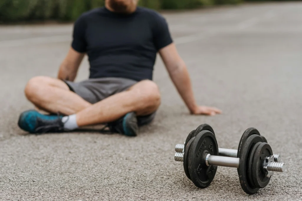 A man sits on the ground in the background, appearing fatigued after a workout, with a heavy dumbbell in the sharp focus of the foreground. This is a visual representation of the physical exhaustion often associated with low testosterone.