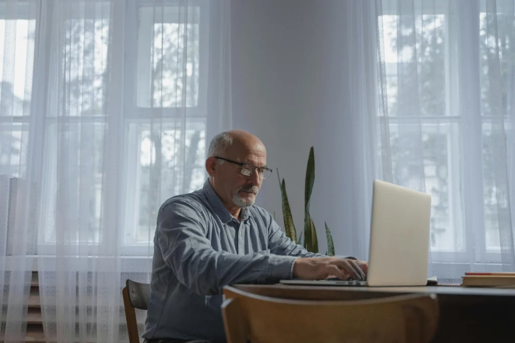 A mature man with a beard and glasses sits at a wooden table, researching Fountain TRT reviews online.