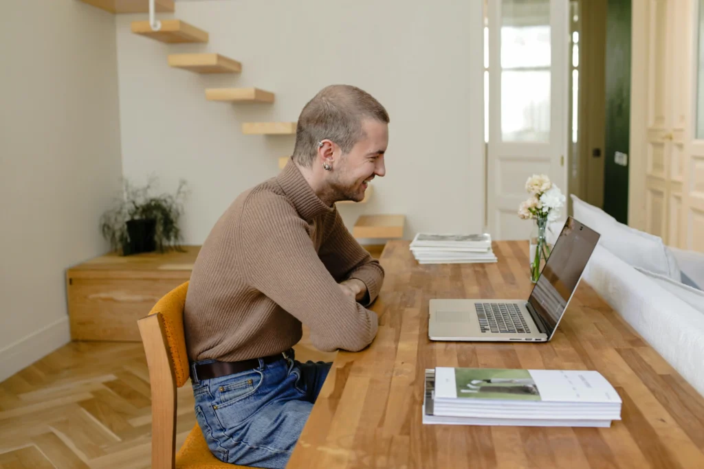 A smiling man sitting at a wooden desk and looking at his laptop, illustrating a patient reviewing his Hone Health TRT cost and membership details during a telehealth consultation.