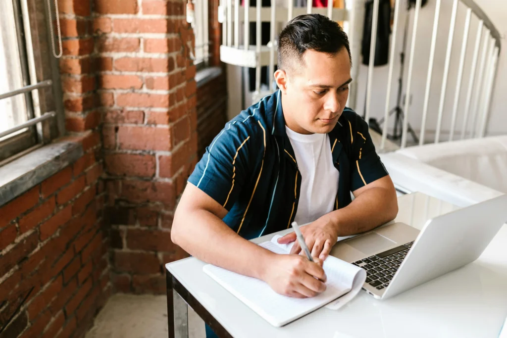 Man writing notes beside a laptop in a modern workspace while researching hormone therapy options and comparing fountain trt vs trt nation online.