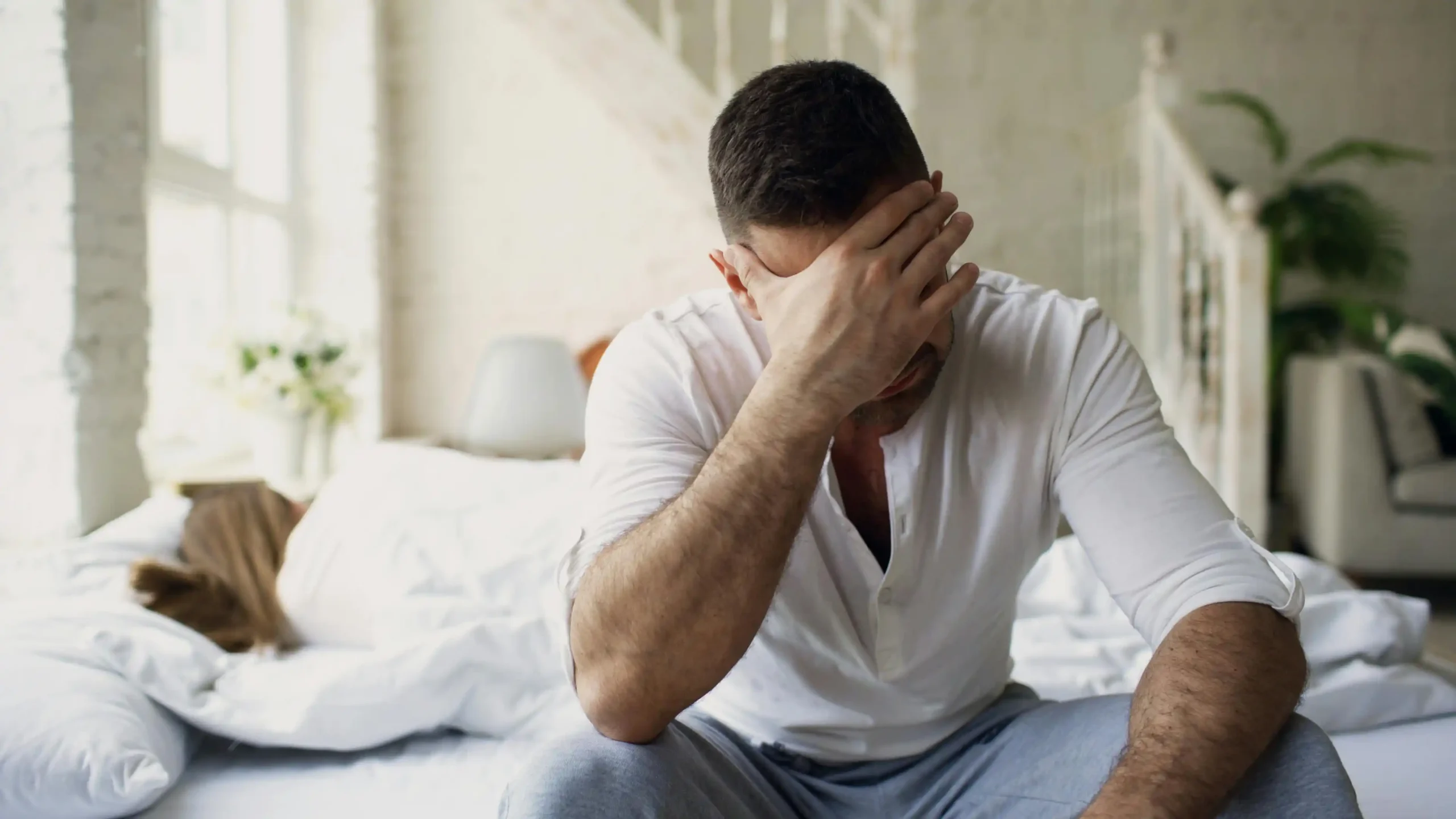 Tired man in his 40s sitting on the edge of a bed in dim morning light, illustrating the fatigue often associated with low testosterone.