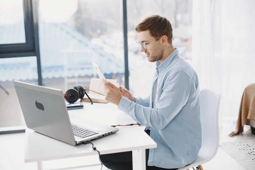Person using a tablet at a home office desk with a laptop and headphones while researching the best ED medication online and telehealth treatment options.