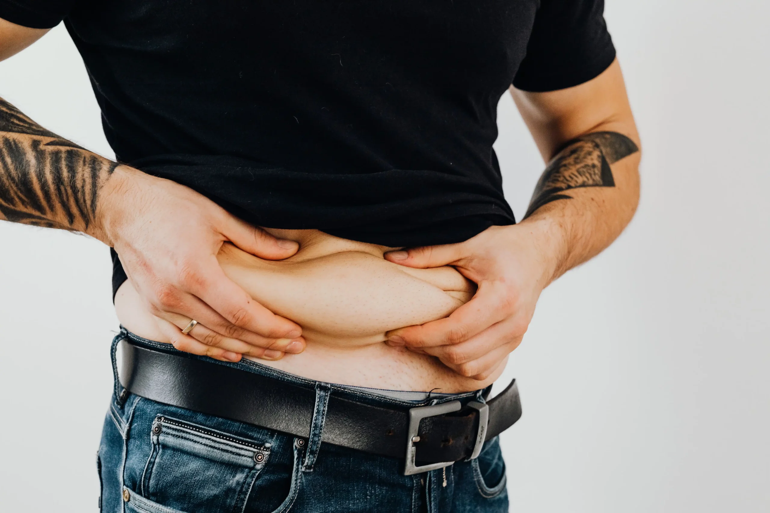 A person wearing a black shirt and jeans pinches excess abdominal fat around their waist, illustrating a weight management topic for compounded semaglutide vs tirzepatide and the comparison of medications used to support weight loss and obesity treatment.