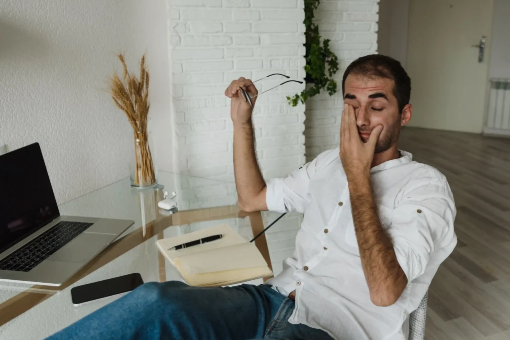 A tired man sits at a glass desk in a home office, holding his glasses while rubbing his eyes beside an open laptop and notebook. His fatigued expression illustrates symptoms commonly associated with hormonal imbalance, making the image suitable for content about estradiol on TRT, estrogen management, and men's hormone health.