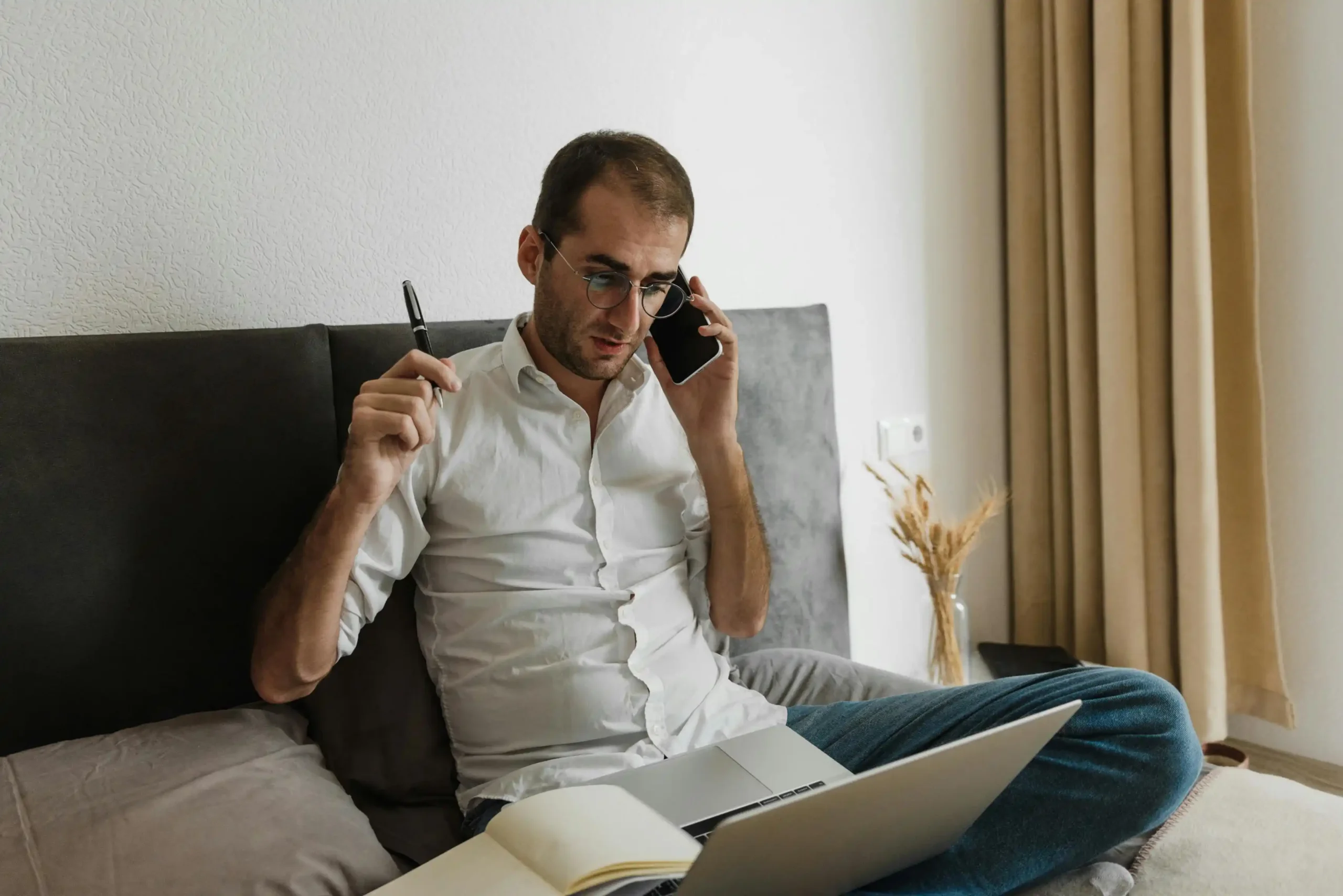 Man comparing Hims vs Roman TRT while taking a phone call and working on a laptop from bed, reviewing notes during a remote consultation at home.