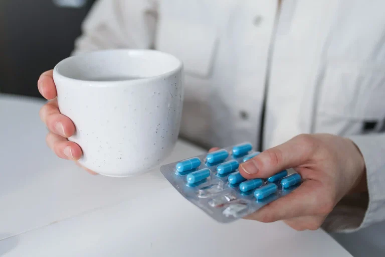 Person holding a blister pack of blue capsules and a mug of water, illustrating medication and treatment options often compared in BlueChew vs Hims discussions about men's health solutions.
