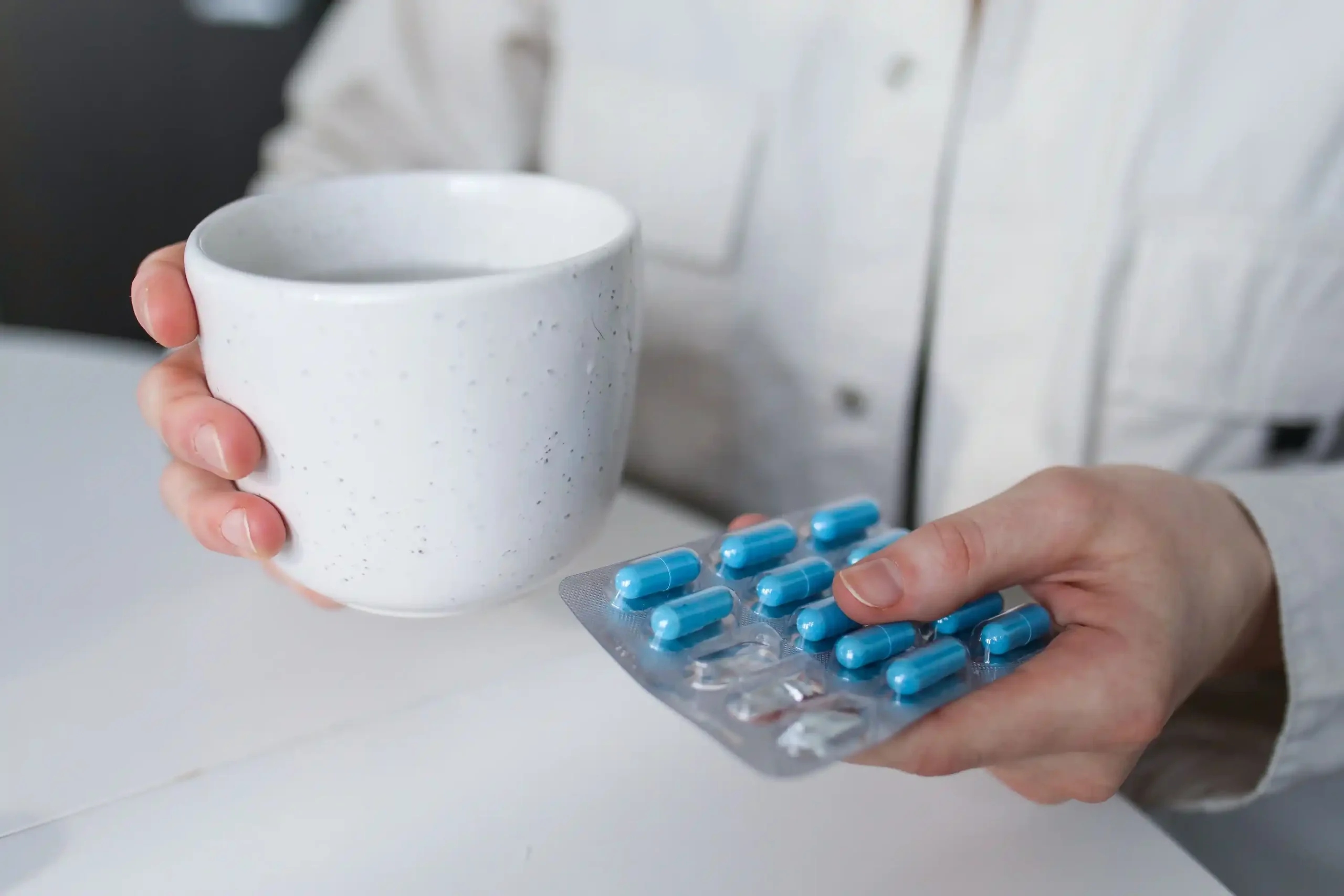 Person holding a blister pack of blue capsules and a mug of water, illustrating medication and treatment options often compared in BlueChew vs Hims discussions about men's health solutions.