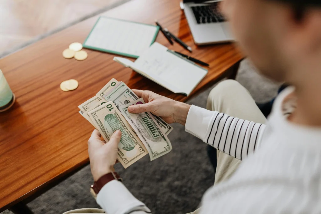 Person counting U.S. dollar bills at a desk with a laptop and financial documents, illustrating cost considerations often discussed in BlueChew vs Hims comparisons for men's health treatments.