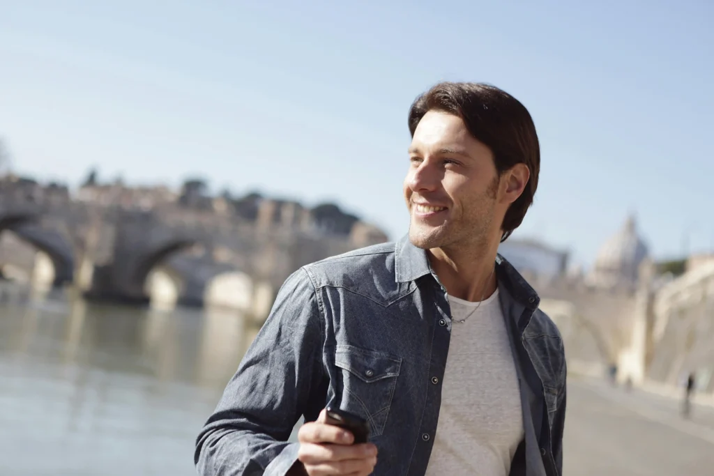 Smiling man standing outdoors near a river with a smartphone in hand, representing confidence and lifestyle benefits often highlighted in BlueChew vs Hims comparisons for men's health and wellness solutions.