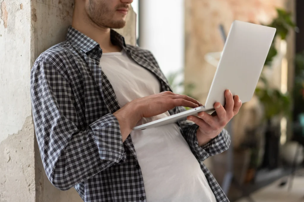 Person leaning against a wall while using a laptop to research treatment options and read a detailed Roman review before making a healthcare decision.