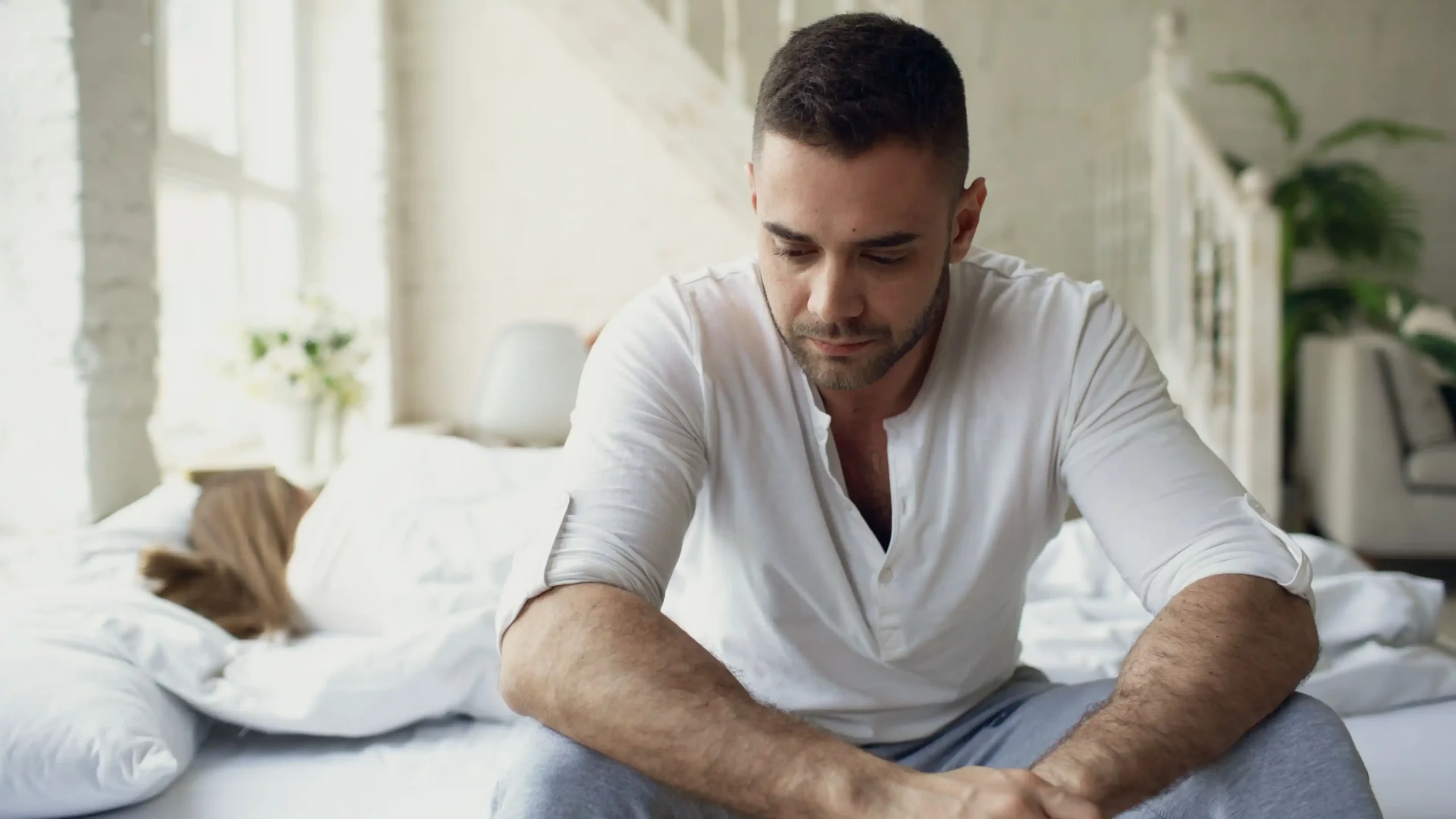 A man sits on the edge of a bed with his head lowered, appearing fatigued and deep in thought, illustrating symptoms commonly discussed in a Rugiet review related to men's sexual health, energy levels, and performance concerns.