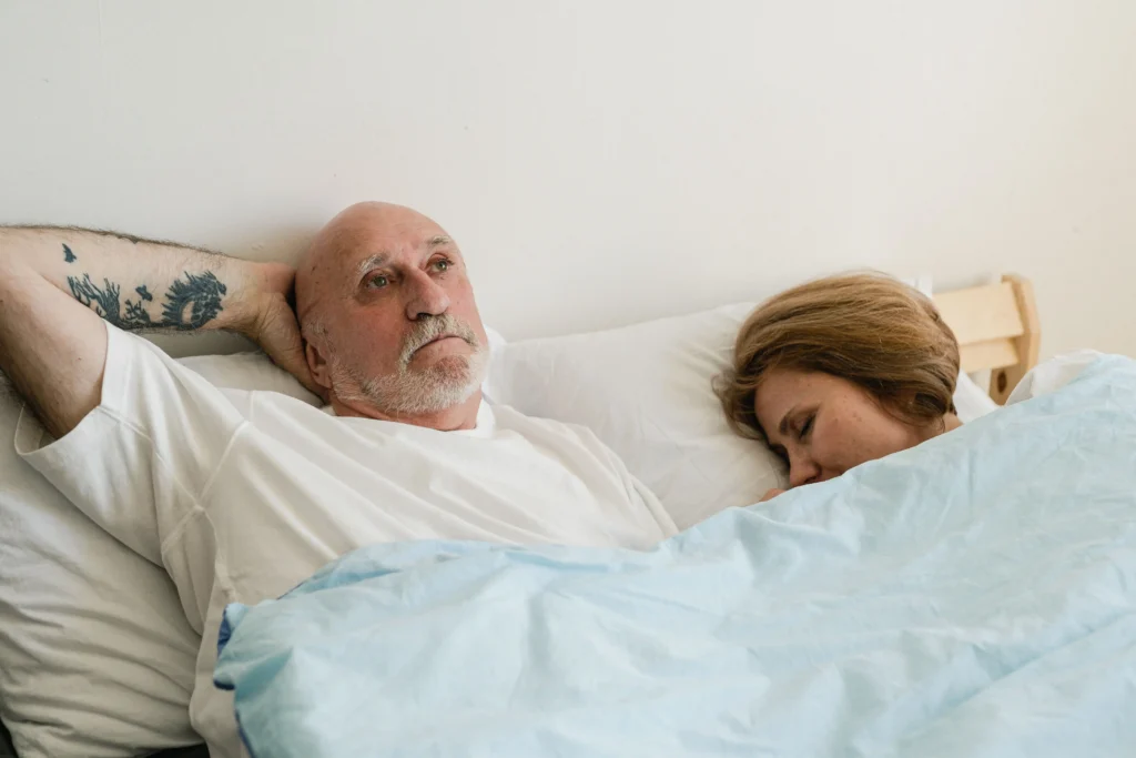 An older man lies awake in bed while his partner sleeps beside him, conveying concerns about erectile dysfunction and intimacy that are commonly compared in a Rugiet vs Hims discussion.