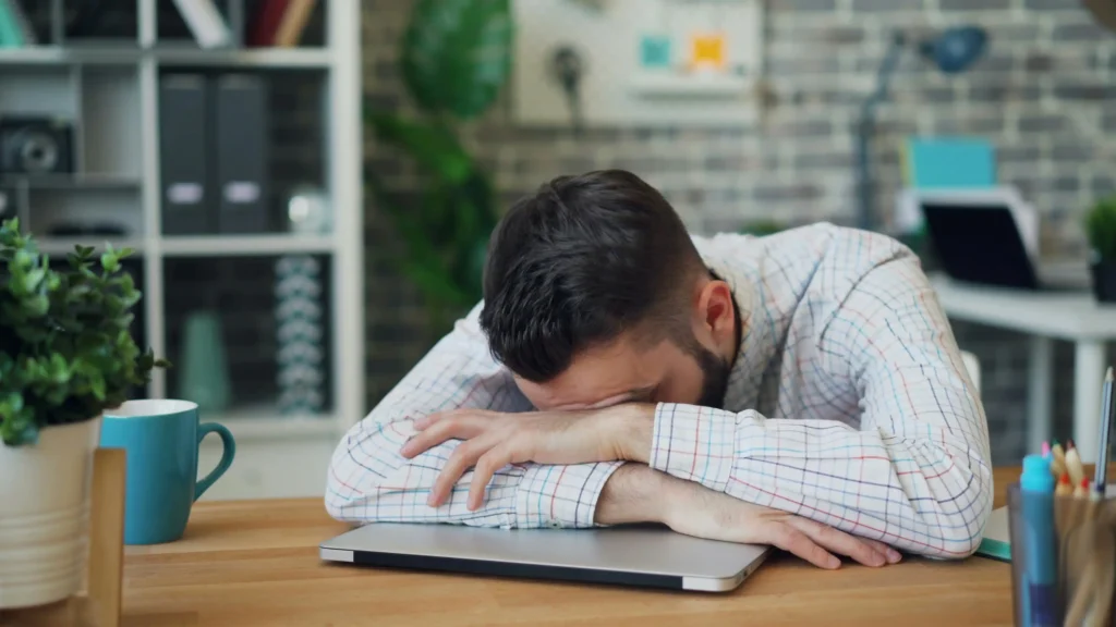 Tired man resting his head on a laptop at work, highlighting fatigue and low energy concerns often discussed in Hims vs Roman TRT comparisons.