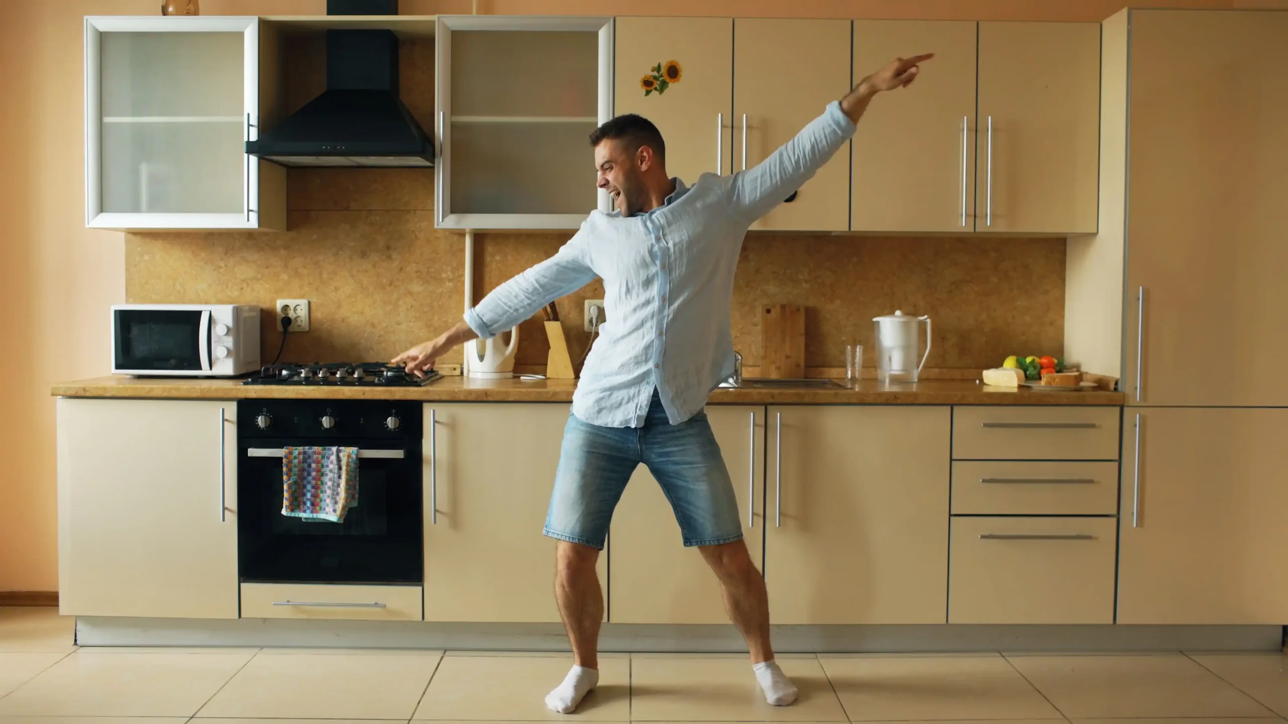 A man dances enthusiastically in a modern kitchen with his arms outstretched, wearing a light blue shirt, denim shorts, and socks. The bright indoor setting conveys energy and vitality, making it suitable for content discussing TRT estrogen, hormone balance, or men's health.