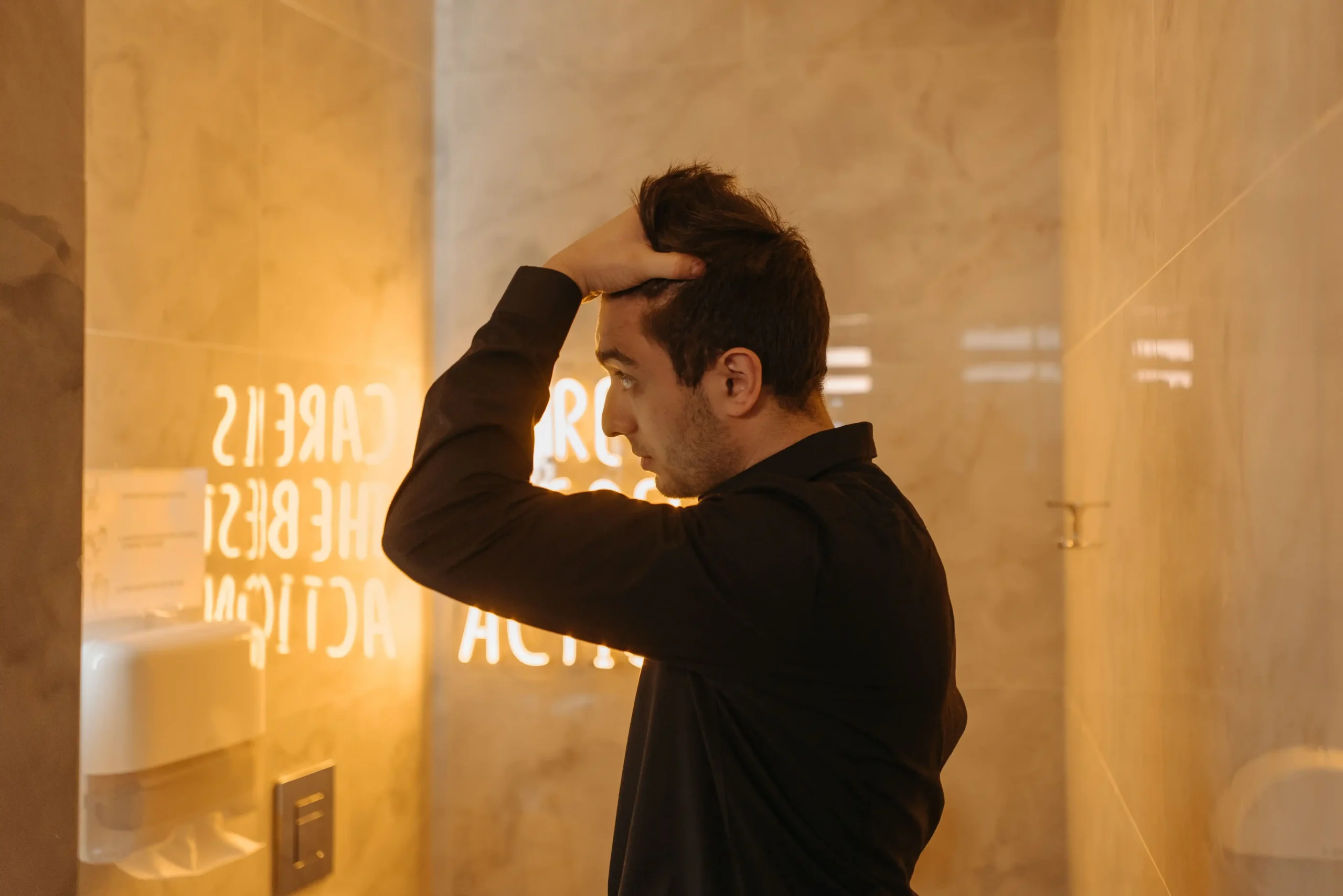 Young man inspecting his hairline in a bathroom mirror for a Hims vs Keeps hair loss treatment comparison.