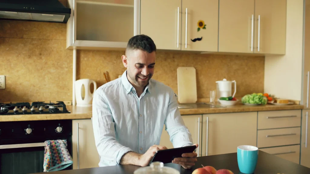 Man smiling as he uses a tablet at his kitchen counter, researching treatment options for hair loss in men through an online consultation.