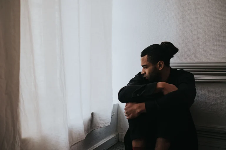 Man sitting alone by a window looking thoughtful, representing the emotional impact of seeking erectile dysfunction treatment.