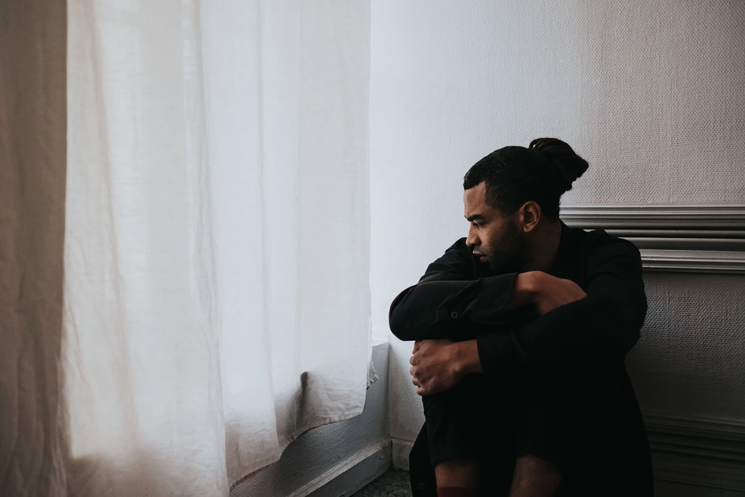 Man sitting alone by a window looking thoughtful, representing the emotional impact of seeking erectile dysfunction treatment.