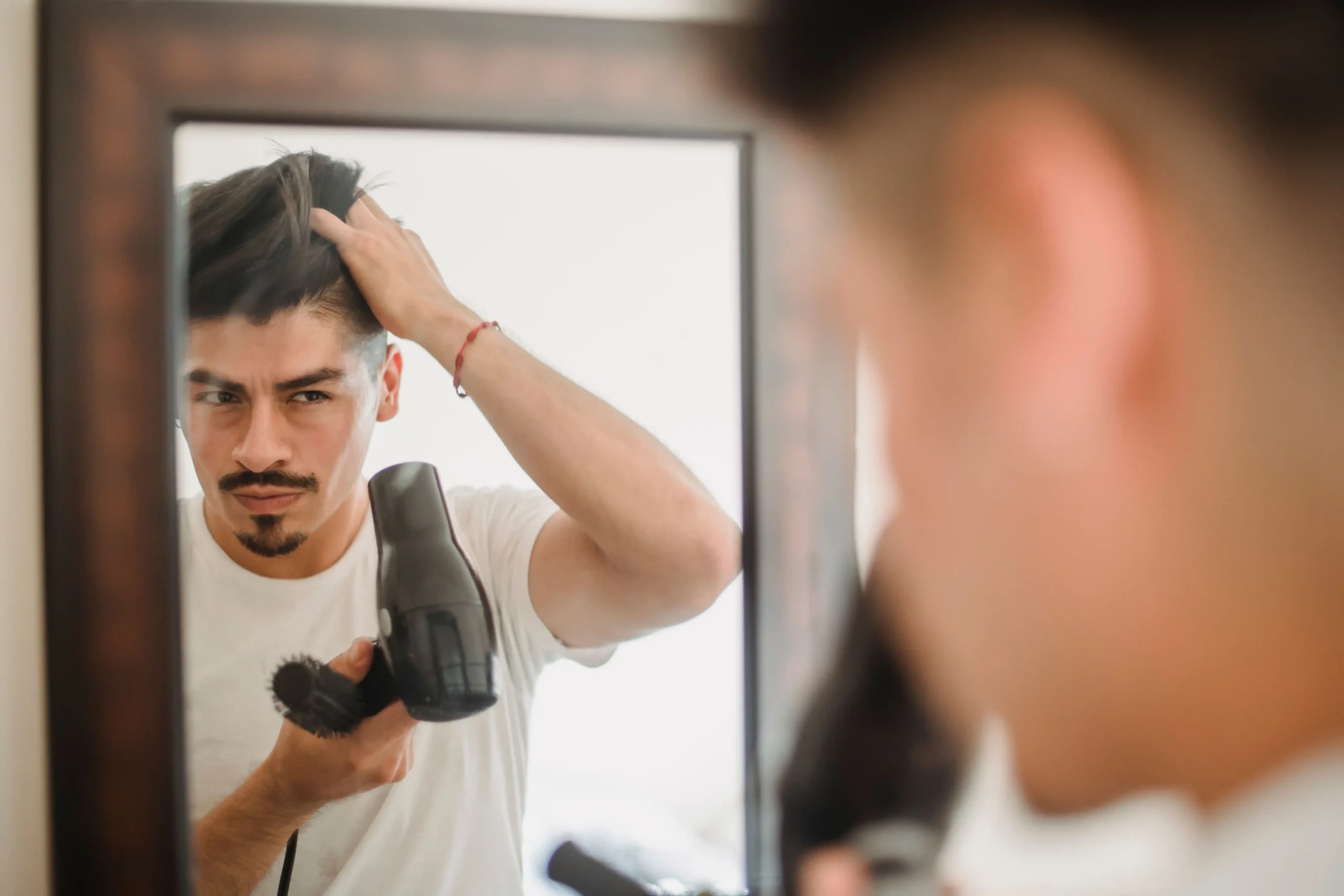 Man in a white t-shirt lifting his hair in front of a bathroom mirror while holding a hairdryer and round brush, checking his hairline for early signs of hair loss in men.
