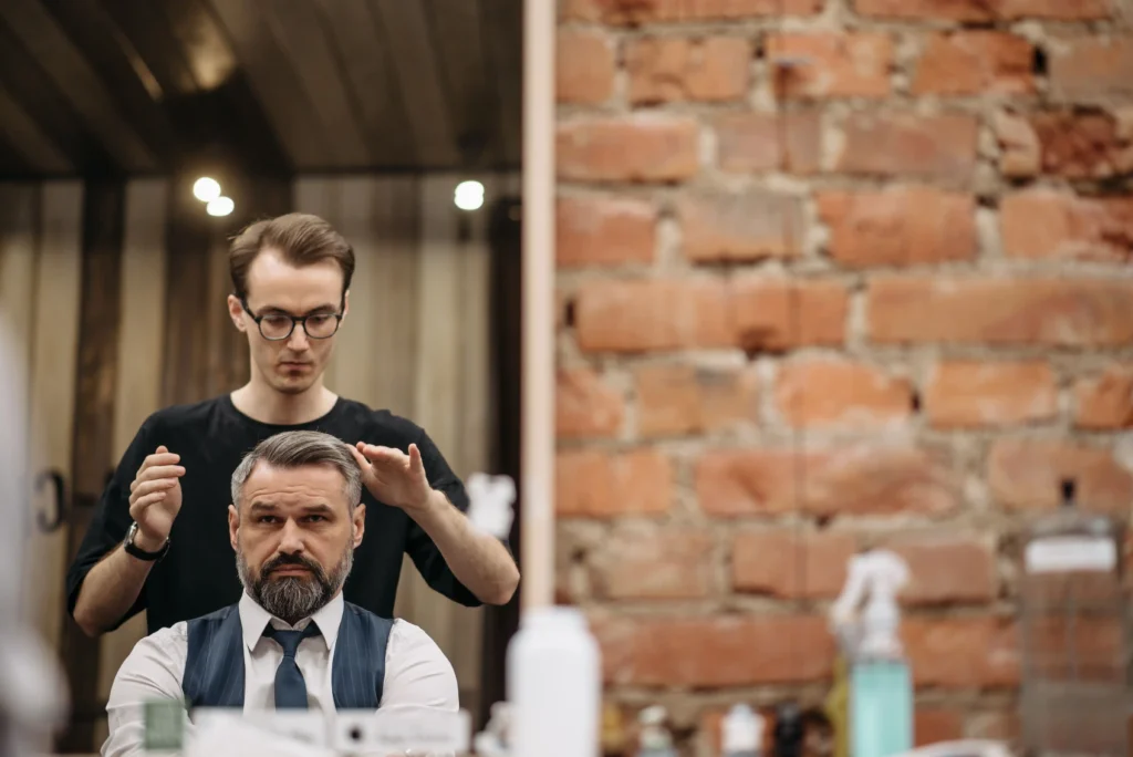 Barber standing behind a seated client in a barbershop chair, lifting and inspecting his greying hair at the temples where male pattern baldness typically begins.