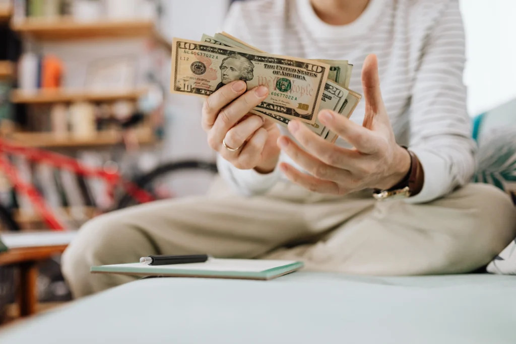 Person counting U.S. dollar bills while considering healthcare costs and a TRT Nation discount code.