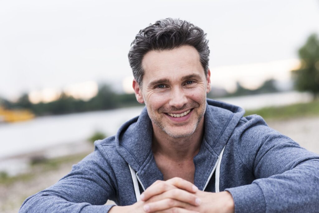A smiling man in his forties in a grey hoodie, outdoors by a river